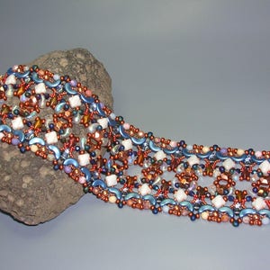 Puede incluir: Un brazalete de cuentas de múltiples hilos con un cierre dorado. El brazalete presenta una variedad de colores, incluyendo azul, marrón, blanco y rosa. Las cuentas están dispuestas en un patrón geométrico.
