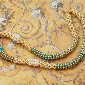 以下が含まれることがあります： 金、白、ターコイズ色のビーズで作られたネックレス。ビーズはらせん状に配置されています。背景はヴィンテージの花柄の壁紙です。美しいジュエリーです。