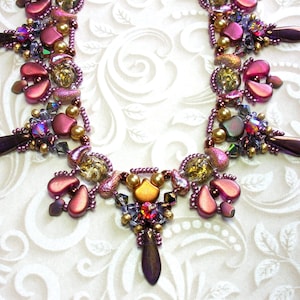 Può includere: Una collana di perline con uno schema di colori viola, oro e iridescente. La collana presenta una varietà di perline, tra cui perline a forma di lacrima, perline rotonde e perline sfaccettate. La collana è progettata in uno schema a cascata.