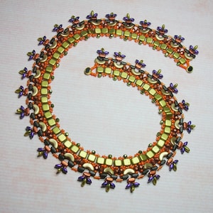 Op de afbeelding: Een kralenketting met een geometrisch ontwerp in tinten groen, oranje en paars. De ketting is gemaakt met kleine, vierkante kralen en heeft een herhalend patroon van bloemen.