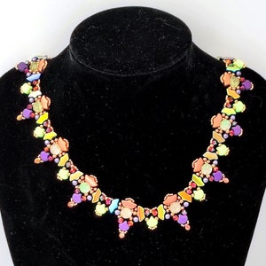 Può includere: Una colorata collana di perline esposta su un manichino nero. La collana presenta una varietà di forme e colori, tra cui viola, verde, arancione e giallo. Le perline sono disposte in un motivo geometrico.