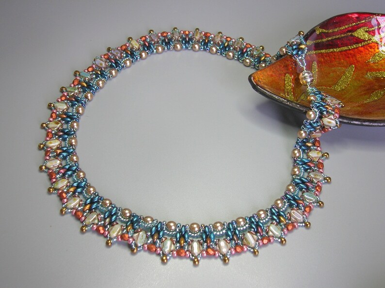 Puede incluir: Un collar de cuentas multicadena con un dise&ntilde;o graduado. El collar presenta una variedad de colores, incluyendo azul verdoso, cobre y crema. Las cuentas est&aacute;n dispuestas en un patr&oacute;n geom&eacute;trico, creando un aspecto &uacute;nico y llamativo.