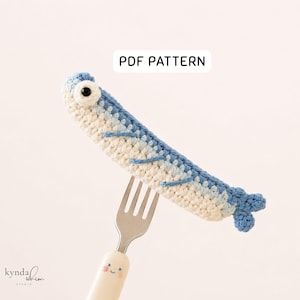 Op de afbeelding: Een gehaakte vis in blauw- en wittinten, rustend op een vork. De vis heeft een groot oog en een kleine staart. De tekst "PDF PATTERN" wordt boven de vis weergegeven. De vork heeft een lachend gezicht op de handgreep.
