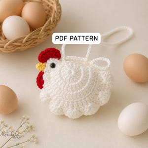 Könnte beinhalten: Ein weißer, gehäkelter Hühnerbeutel mit roten und gelben Akzenten, einem schwarzen Auge und einer weißen Schlaufe. Der Text "PDF PATTERN" ist zu sehen. Der Beutel ist von Eiern in einem Korb und auf einer hellen Oberfläche umgeben.
