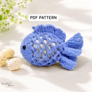 May include: A blue crocheted fish cover with a lattice pattern, featuring a small black eye and a textured fin. The item is labeled PDF PATTERN. Two speckled cream-colored eggs are positioned to the left of the blue crocheted fish figure.