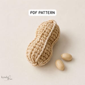 Puede incluir: Una cáscara de cacahuete de ganchillo en color beige con una superficie texturizada y dos pequeñas cuentas ovaladas lisas de color claro colocadas al lado. La imagen incluye el texto PDF PATTERN arriba y la marca kynda whim studio abajo.