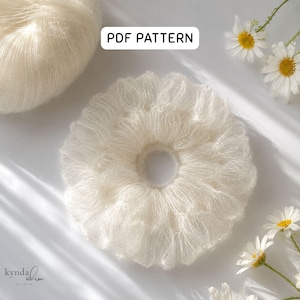 May include: A fluffy, off-white crocheted scrunchie with a central hole, next to a ball of yarn. The image includes the text "PDF PATTERN" and the logo "kynda whim studio". Daisies are scattered around the scrunchie.
