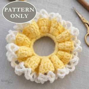 Puede incluir: Una goma para el pelo hecha a mano, de ganchillo amarillo y blanco, que se asemeja a un girasol. El accesorio para el cabello presenta una abertura central y está adornado con un borde blanco con volantes. El texto "PATTERN ONLY" está en un círculo gris.