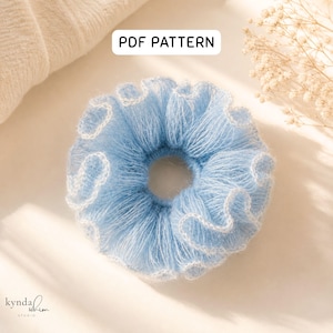 Puede incluir: Una scrunchie azul claro con una textura esponjosa y ribete blanco. La scrunchie es redonda con un agujero en el centro. El texto "PDF PATTERN" está en un rectángulo blanco. La scrunchie está sobre un fondo claro.