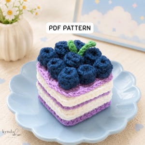 Könnte beinhalten: Ein quadratischer gehäkelter Kuchen mit Schichten aus weißem und lila Garn, belegt mit dunkelblauen gehäkelten Blaubeeren und grünen Blättern. Der Kuchen steht auf einem hellblauen, gewellten Teller. Der Text "PDF PATTERN" befindet sich in einem weißen Rechteck.