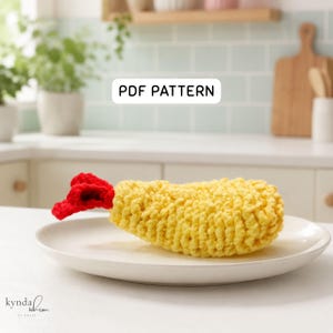 Può includere: Un modello all'uncinetto per gamberi in tempura con l'etichetta PDF PATTERN. L'oggetto è una forma di gambero fritto giallo e testurizzato con una coda rosso brillante, appoggiato su un piatto bianco. Il design imita un cibo fritto con maglie.