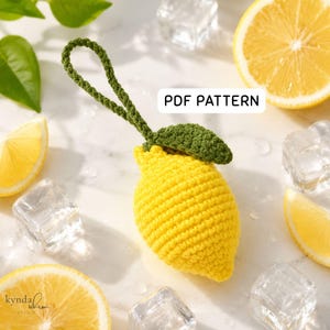 Può includere: Un limone all'uncinetto giallo con una foglia verde e un occhiello per appenderlo. Un'etichetta rettangolare bianca con il testo nero PDF PATTERN è visibile. Il limone è circondato da fette di limone e cubetti di ghiaccio su una base bianca.