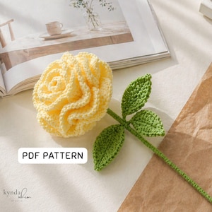 May include: A crocheted rose in pale yellow, featuring green leaves and a stem. The flower has detailed, textured petals. The image includes the text "PDF PATTERN" and the studio's logo.