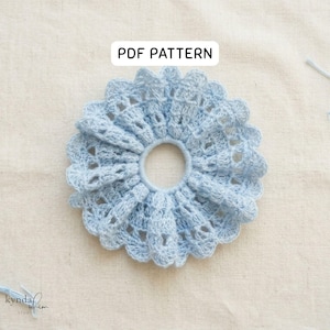 May include: A light blue crocheted scrunchie with a ruffled edge and openwork design. The scrunchie is centered on a neutral fabric background. Text on the image reads "PDF PATTERN".