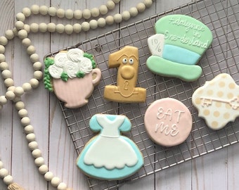 May include: A set of six Alice in Wonderland themed sugar cookies. The cookies are decorated with various characters and objects from the story, including a teacup with flowers, a number one, the Mad Hatter's hat, a dress, a key, and a cookie that says "Eat Me".