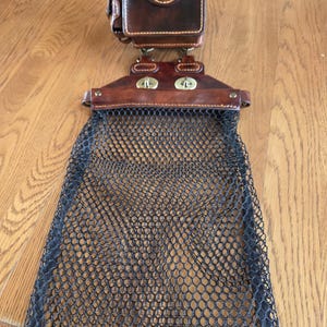 May include: A brown leather fishing creel with a black mesh net. The creel has a small, square compartment with a decorative brass button and two brass clasps. The net is attached to the creel.