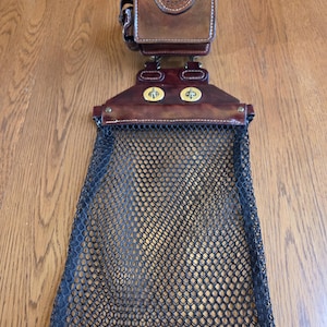 May include: A brown leather fishing creel with a black mesh net. The creel has a rectangular top with a decorative flap and a gold-colored clasp. The net is attached to the bottom of the creel.
