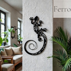 Puede incluir: Arte de pared de metal negro con forma de gecko con un diseño decorativo y una cola en espiral. El gecko está montado en una pared blanca texturizada. La palabra "Ferro" es visible.
