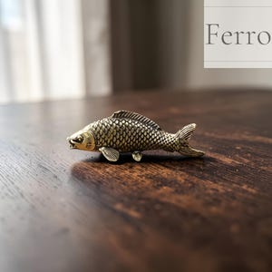 Puede incluir: Una detallada figura de pez de latón con escamas y aletas texturizadas. El pez está colocado sobre una superficie de madera oscura, con la palabra "Ferro" visible en el fondo. La figura tiene un diseño realista y un acabado metálico.