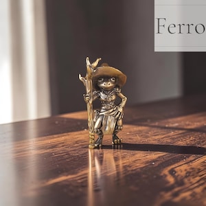 May include: A detailed bronze figurine of a frog dressed in a hat and holding a staff. The frog is wearing a hat and a vest, with a belt around its waist. The figurine is standing on a wooden surface. The word "Ferro" is in the background.