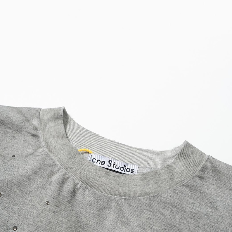 May include: A heather gray crew neck sweatshirt featuring small metallic studs scattered across the fabric. The interior collar displays a white brand label with the text Acne Studios in black, accompanied by a small yellow fabric loop.
