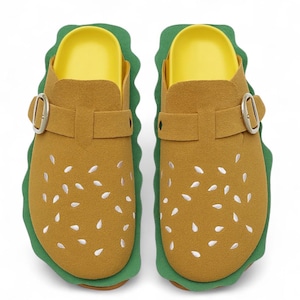 Könnte beinhalten: Ein Paar ausgefallene Clogs, die wie Hamburger aussehen sollen. Die Schuhe haben ein braunes Wildleder-ähnliches Material mit weißen Samendetails, eine grüne, salatartige Einfassung und eine gelbe Innensohle. Jeder Clog hat eine silberne Schnalle und einen Riemen.