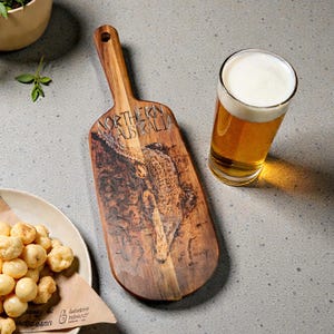 May include: Wooden serving board with a handle, featuring a crocodile illustration and the text "NORTHERN AUSTRALIA." A glass of beer and a plate of snacks are also present. The board is approximately 15 inches long.