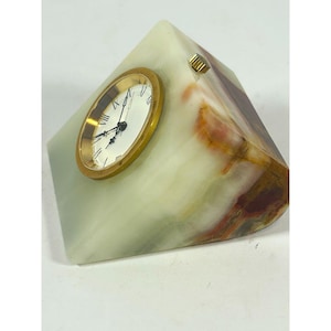 May include: A wedge-shaped desk clock crafted from polished light green and brown banded stone. The clock face is circular with a gold-toned metal bezel, black hands, and black Roman numerals. A small gold-toned adjustment knob is visible on the top.
