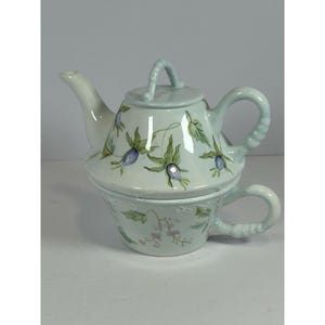 May include: A light blue ceramic teapot and teacup set. The teapot has a lid and a handle, and the teacup is stacked underneath. Both pieces are decorated with green leaves and blue and purple flowers.