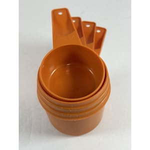 Puede incluir: Un juego de cuatro tazas medidoras de plástico naranja encajables con mangos largos, cada una con un pequeño orificio circular en el extremo para colgar. Las tazas están apiladas verticalmente y presentan un acabado mate uniforme.