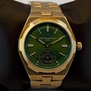 Puede incluir: Un reloj de pulsera dorado con esfera verde. El reloj tiene una esfera redonda con manecillas y marcadores dorados, y una pequeña subesfera. La correa está hecha de eslabones de metal dorado. El reloj se muestra sobre una superficie oscura.