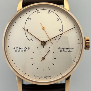 May include: A gold-tone wristwatch with a cream-coloured dial, featuring the text NOMOS GLASHÜTTE, Gangreserve 48 Stunden, and Made in Germany. The watch has thin gold hands, a power reserve indicator sub-dial, a small seconds sub-dial, and a dark brown leather strap.