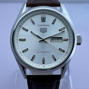 Op de afbeelding: Zilveren Carrera automatisch horloge met een witte wijzerplaat, zwarte wijzers en een datumweergave. Het horloge heeft een bruine leren band en een zilveren kast. De wijzerplaat toont de dag van de week als "MA 7".