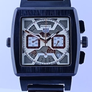 Peut inclure: Une montre TAG Heuer Monaco noire avec un cadran carré. Le cadran est un mélange de beige, de gris et de rouge avec plusieurs cadrans et le mot "MONACO" affiché. La montre a un bracelet en métal noir.