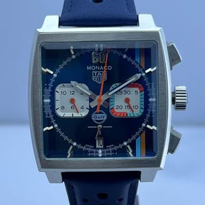 May include: A square-faced wristwatch with a dark blue dial, white and orange accents, and a blue leather strap. The watch face displays the text "MONACO TAG HEUER" and "Gulf". The watch has a silver-coloured case and a blue strap.
