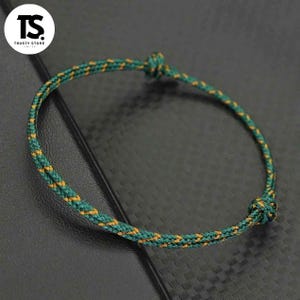 Op de afbeelding: Een groen en geel gevlochten armband met een knoopsluiting. De armband is gemaakt van een geweven materiaal en wordt getoond op een donkere ondergrond. Het logo "TS TRUSTY STORE LIMITED" staat linksboven.