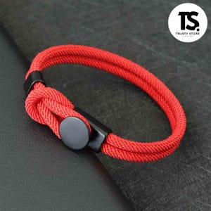 May include: A red rope bracelet with a black clasp and a circular black button. The bracelet is knotted at one end and has a double-strand design. The image includes the text "TRUSTY STORE LIMITED".