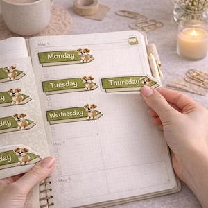 Könnte beinhalten: Ein Wochenplaner mit Spiralbindung, geöffnet auf einer Doppelseite mit Aufklebern eines Cartoon-Hundes. Die Wochentage sind beschriftet. Eine Tasse Kaffee, eine Kerze und Schreibwaren befinden sich im Hintergrund.