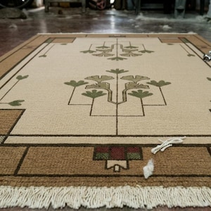 May include: A rectangular beige rug with a geometric design. The rug features a central floral pattern in shades of green and brown, framed by a border of brown and beige rectangles. The rug has a fringe along the bottom edge.