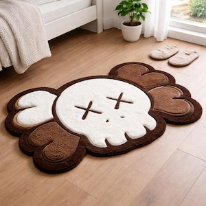 A brown, white, and tan skull-shaped rug with cross-shaped eyes. The rug has a fluffy texture and is placed on a wooden floor. The rug is a decorative item for a bedroom or living space.