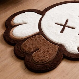 A brown, white, and tan plush rug shaped like a cartoon character's head. The rug features a white face with brown cross-shaped eyes and brown ears. It is placed on a wooden floor.