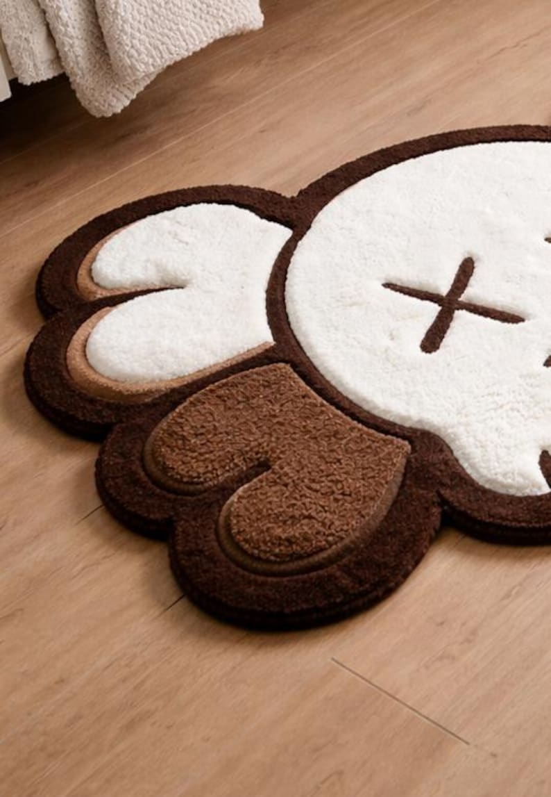 A brown, white, and tan plush rug shaped like a cartoon character's head. The rug features a white face with brown cross-shaped eyes and brown ears. It is placed on a wooden floor.