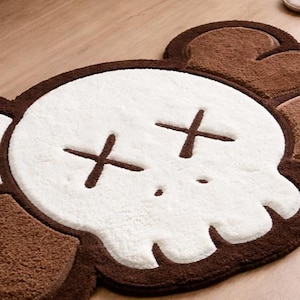 A soft, brown and white skull-shaped rug featuring cross-shaped eyes and a small dot for a nose. The rug has a textured surface and a brown border. This decorative rug is suitable for a bedroom or bathroom.