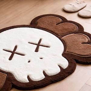 A fluffy, skull-shaped rug featuring a white face and brown crossbones. The rug has a brown border and is on a light wooden floor. Two pairs of beige slippers are in the background.