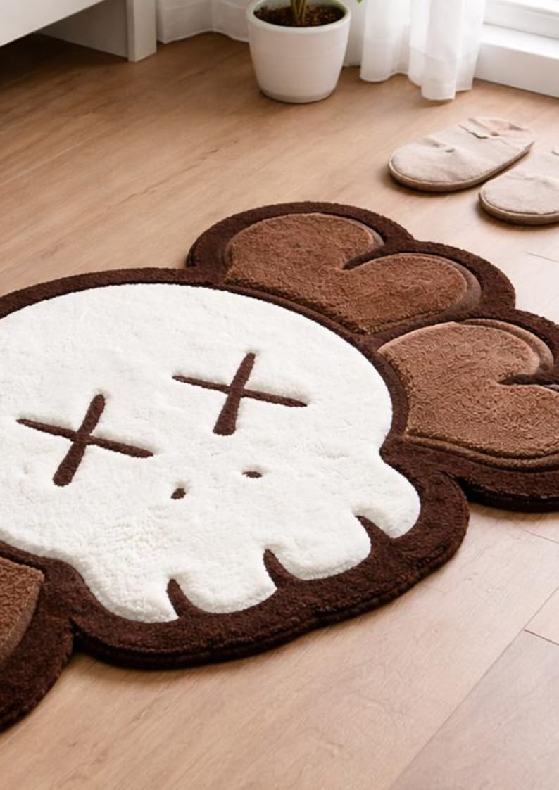 A fluffy, skull-shaped rug featuring a white face and brown crossbones. The rug has a brown border and is on a light wooden floor. Two pairs of beige slippers are in the background.