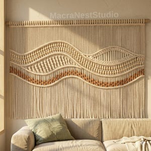 Puede incluir: Un gran tapiz de macramé beige con un diseño ondulado. La pieza presenta intrincados patrones de nudos, detalles de madera y una apariencia texturizada. La decoración de la pared está suspendida de una barra de madera, creando una estética bohemia.
