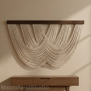 Op de afbeelding: Een macramé wandkleed met een donkerbruine houten balk aan de bovenkant. De crèmekleurige koorden hangen in een vloeiend, symmetrisch patroon naar beneden. Gemonteerd op een lichtbeige muur, boven een houten console.