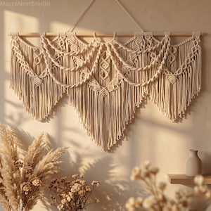 Puede incluir: Un tapiz de pared de macramé beige con una clavija de madera. El intrincado diseño presenta patrones anudados y flecos en cascada. La decoración de la pared está suspendida de un cordón blanco y se exhibe contra una pared de tono neutro. El logotipo de MacraNestStudio está en la esquina superior izquierda.