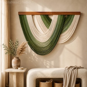 May include: A modern wall hanging with a wooden top and cascading strands in shades of green and cream. The artwork is displayed against a neutral wall, creating a focal point in a contemporary living space. The piece adds texture and color.