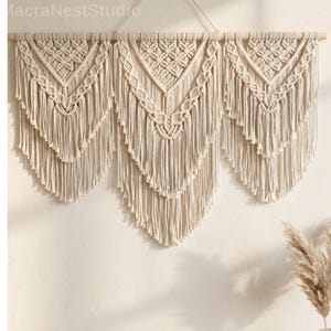 May include: A cream-colored macrame wall hanging with three panels. Each panel features a geometric design at the top and cascading fringe. The piece is suspended from a natural wooden dowel, creating a bohemian aesthetic.